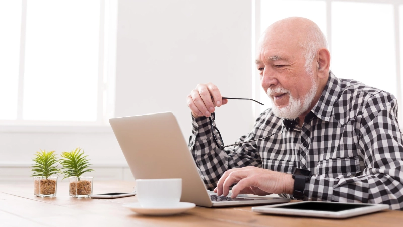 Une personne âgée est assise à table et regarde son ordinateur.