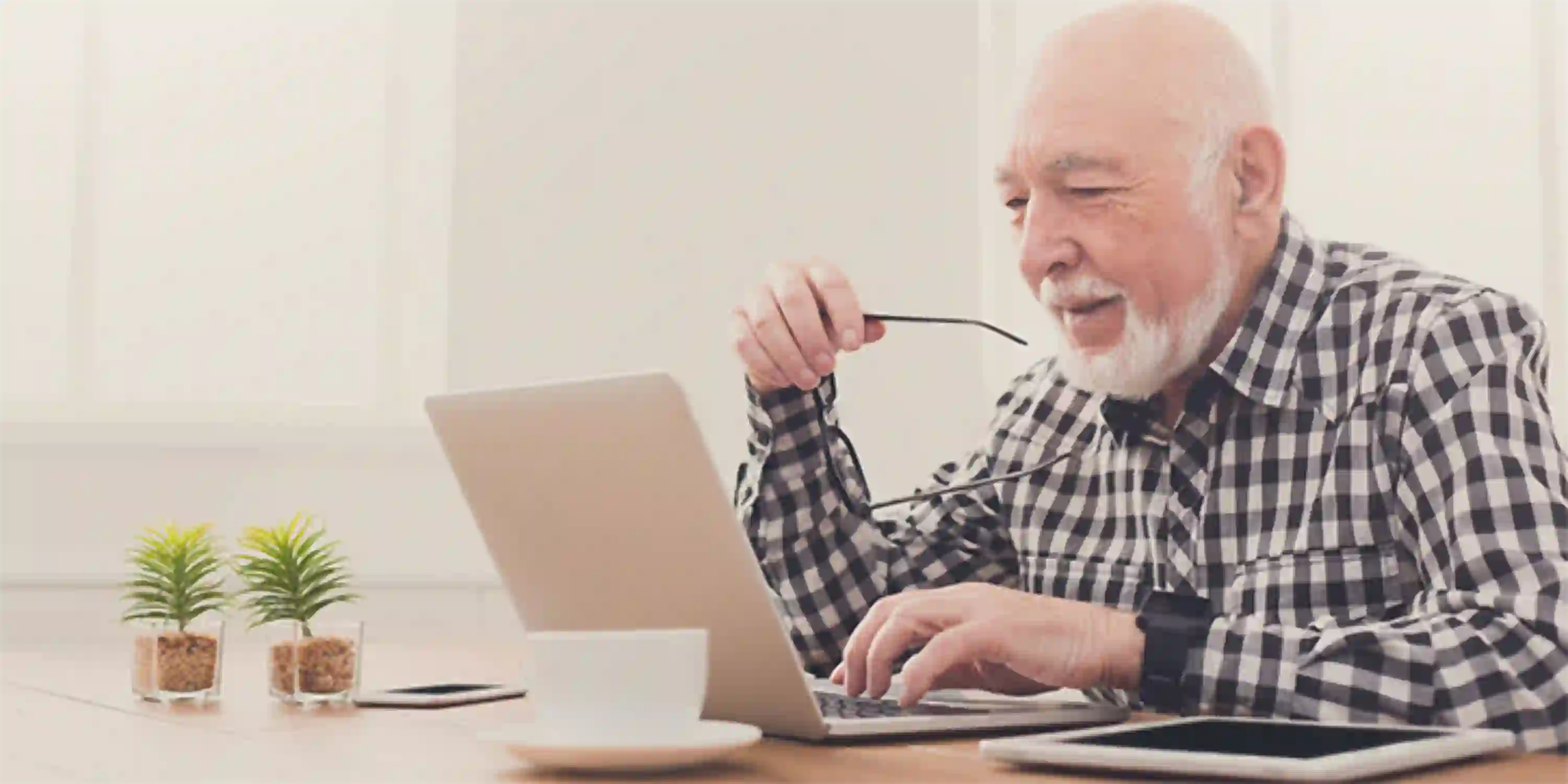 Ein Senior hält seine Brille in der Hand und bedient mit einem Lächeln den Laptop.