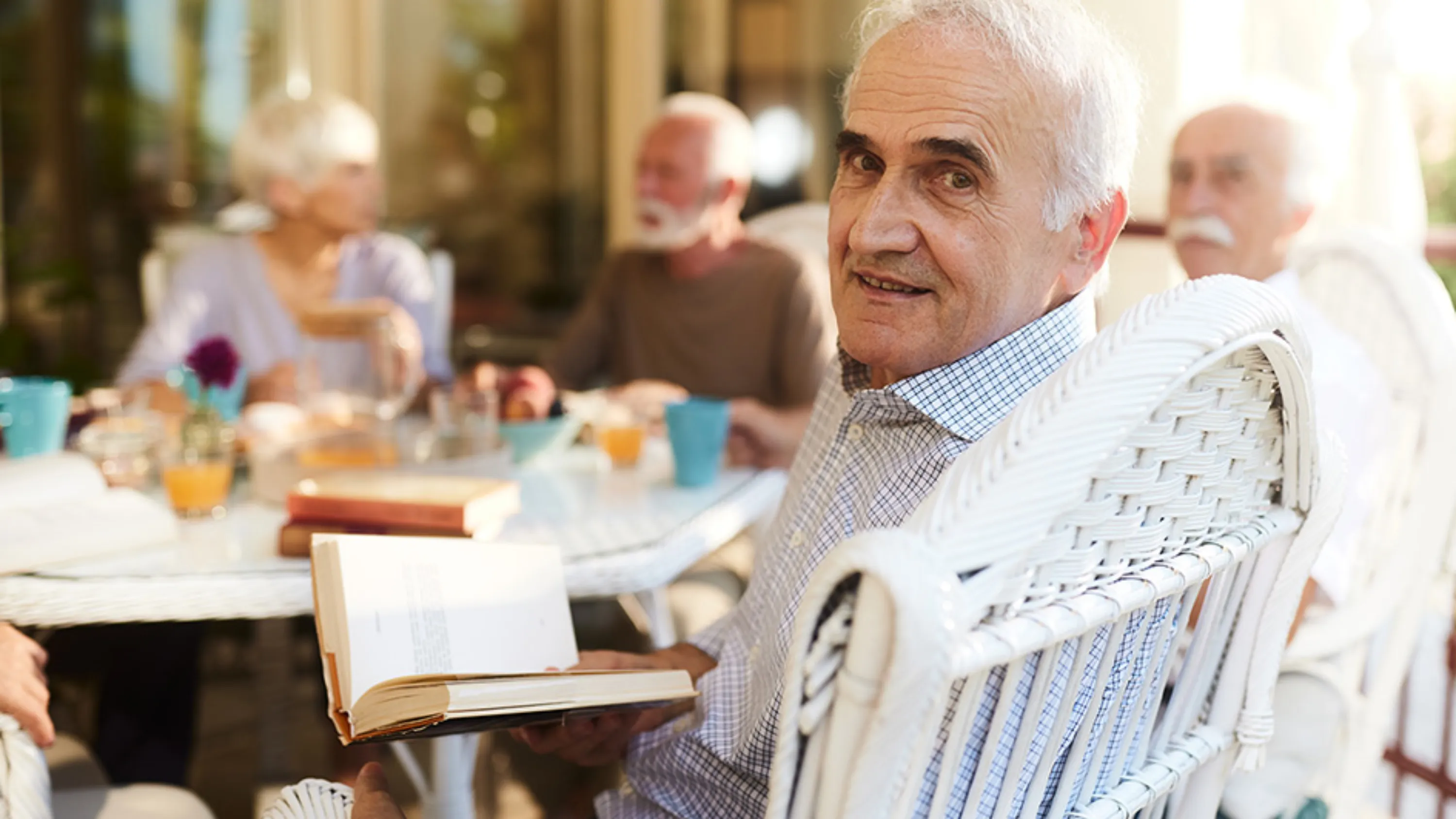 ältere Menschen sitzen an rundem Tisch, Mann schaut in Kamera und hält Buch in der Hand