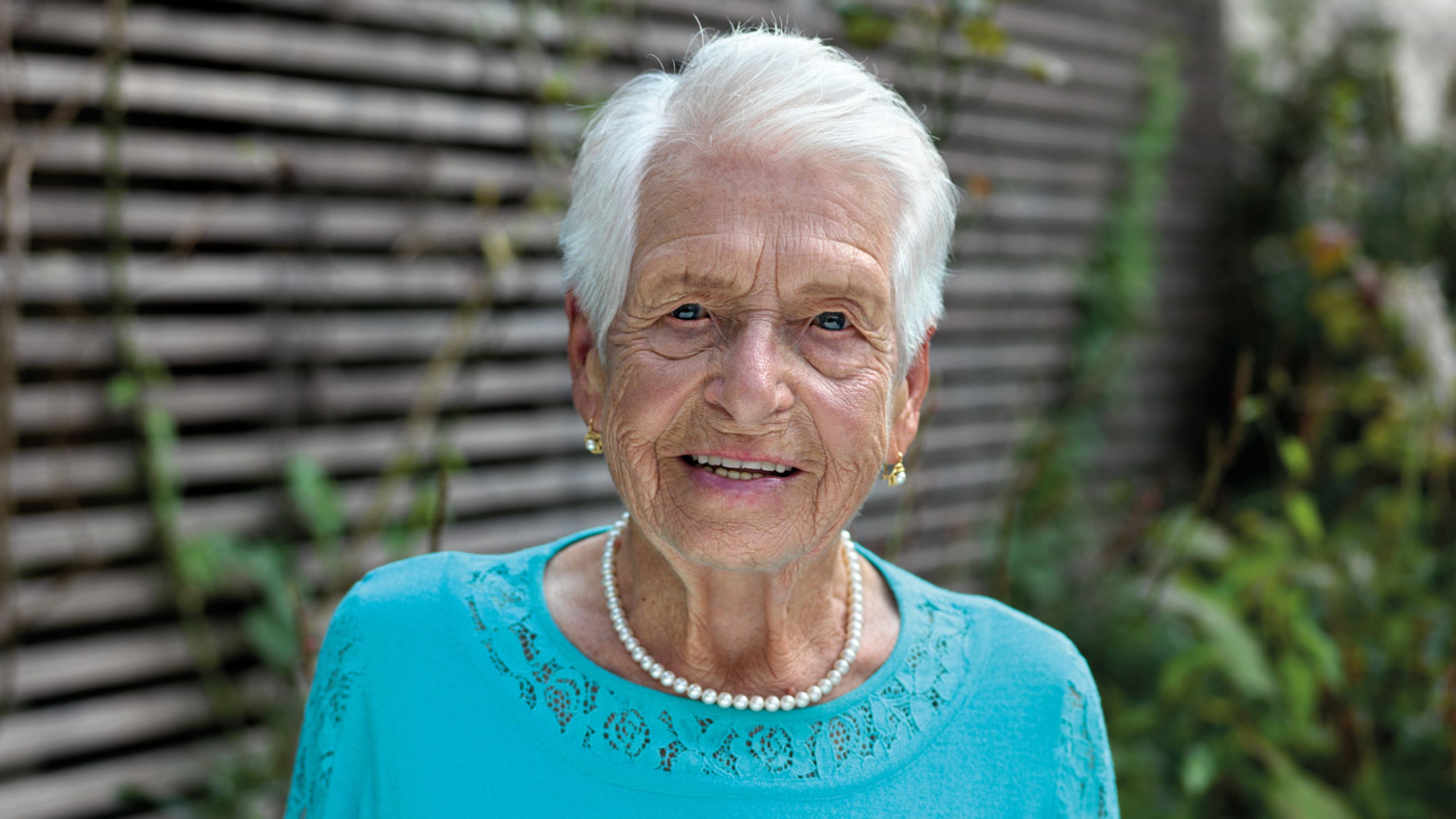 Eine Seniorin mit blauem Top und Perlenkette schaut lächelnd in die Kamera.