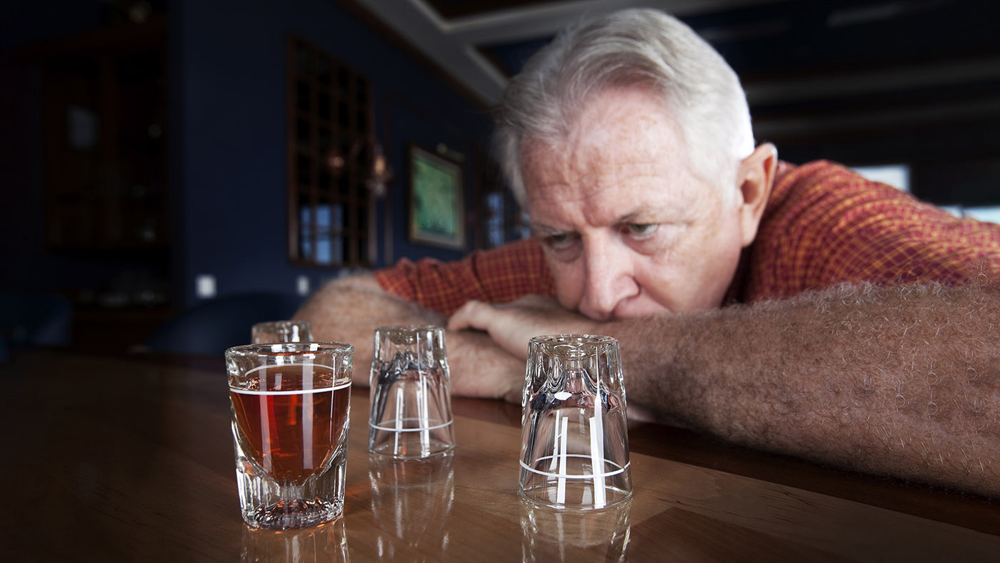 Ein älterer Mann sitzt an der Bar und schaut auf leere und volle Shot-Gläser mit Alkohol.