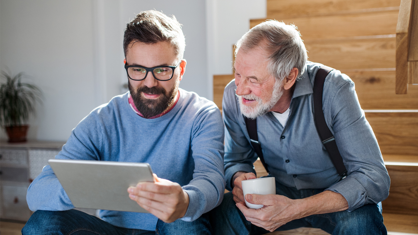 Un homme âgé, une tasse de café à la main, s'informe sur le Docupass avec son fils sur la tablette.