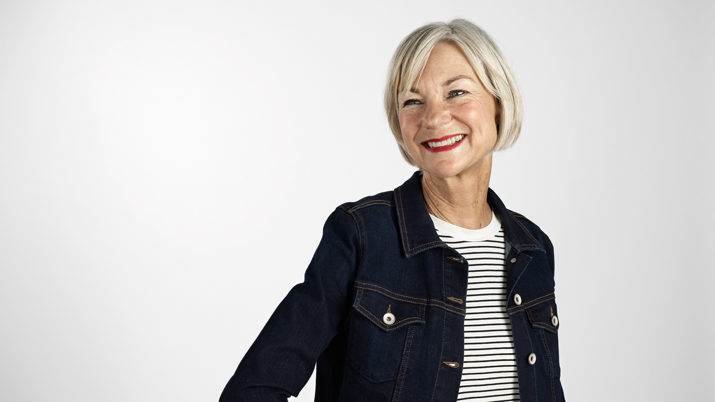 Une femme âgée et souriante aux cheveux gris et à la veste en jean noir.