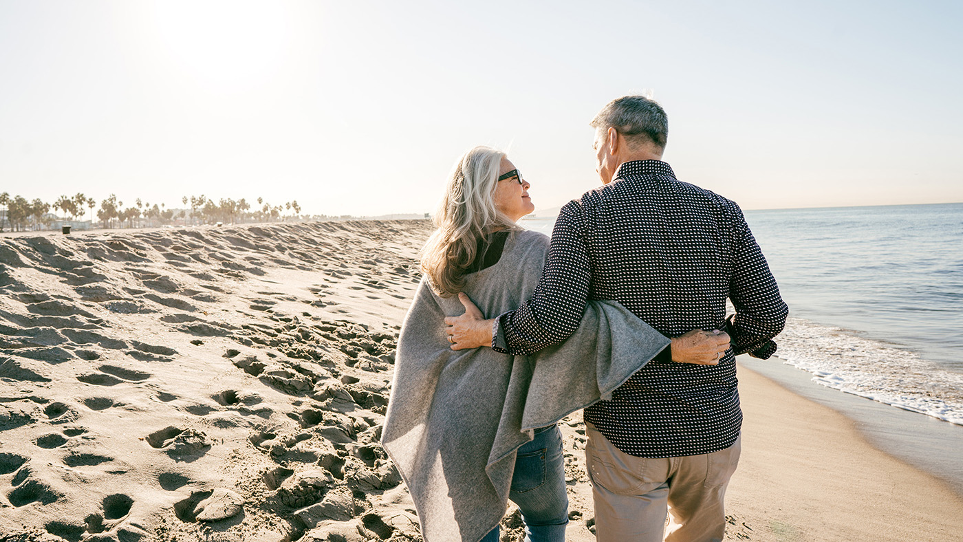 Ein älteres Paar schreitet Arm in Arm am Meer entlang über einen Sandstrand.