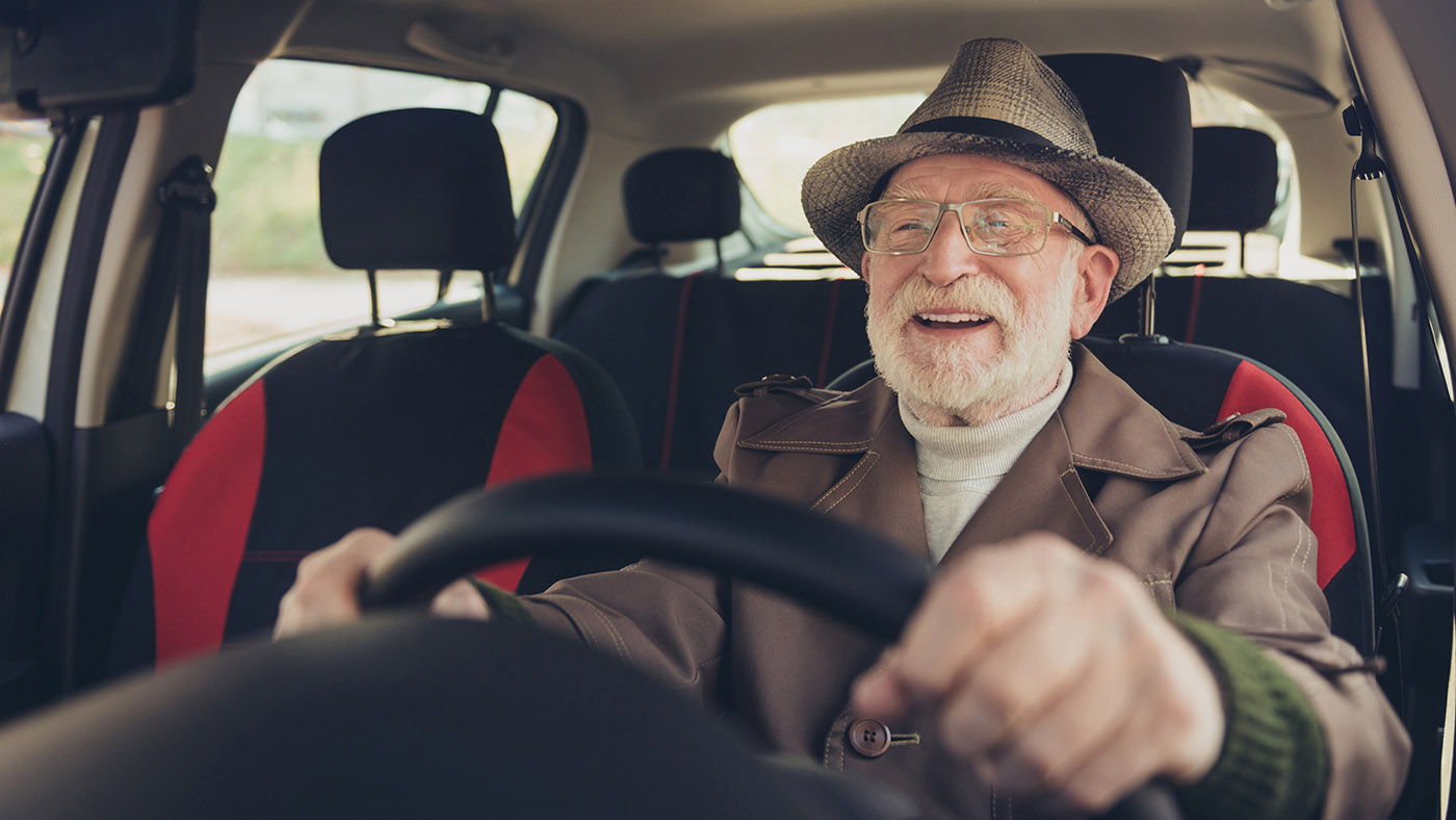 Ein älterer Mann mit Brille, Hut und Bart sitzt am Lenkrad und lächelt.