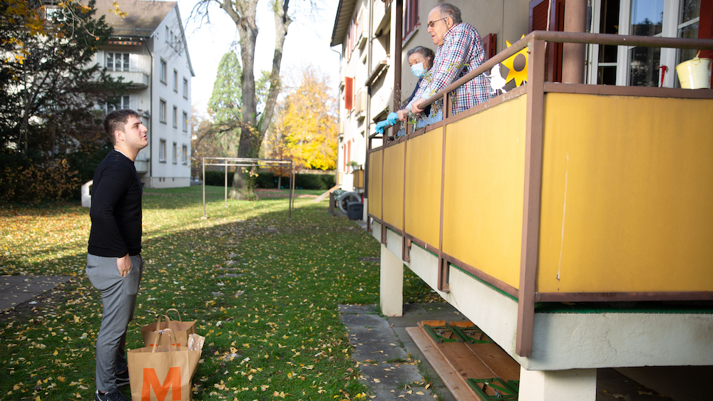 Ein junger Mann steht vor dem Balkon seiner Grosseltern mit zwei vollen Migros Säcken.