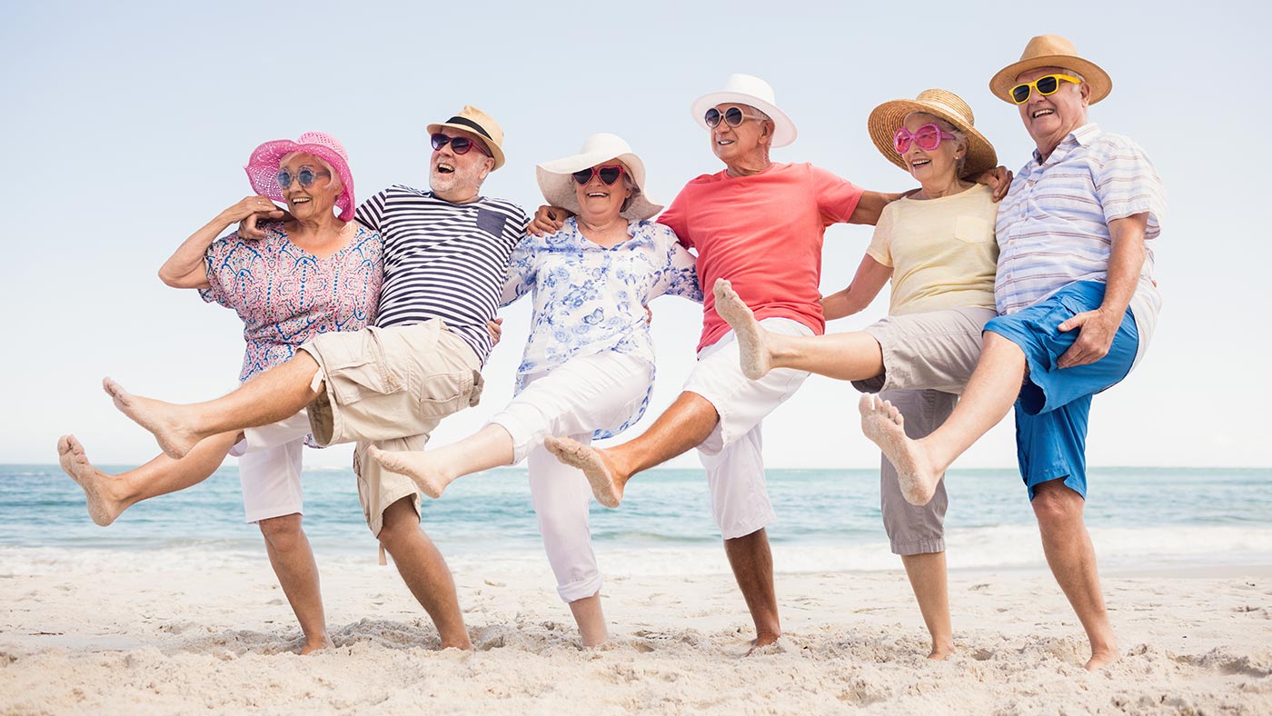 Sechs fröhliche ältere Frauen und Männer mit Hüten und Sonnenbrillen stehen in einer Reihe am Strand und heben tanzend und lachend ein Bein.