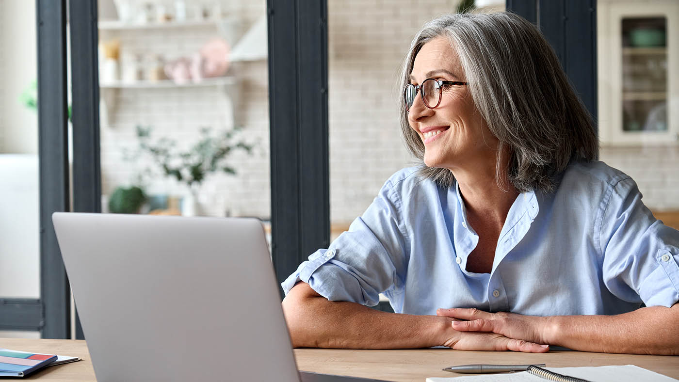 Una donna anziana davanti al computer portatile guarda fuori dalla finestra con un sorriso.