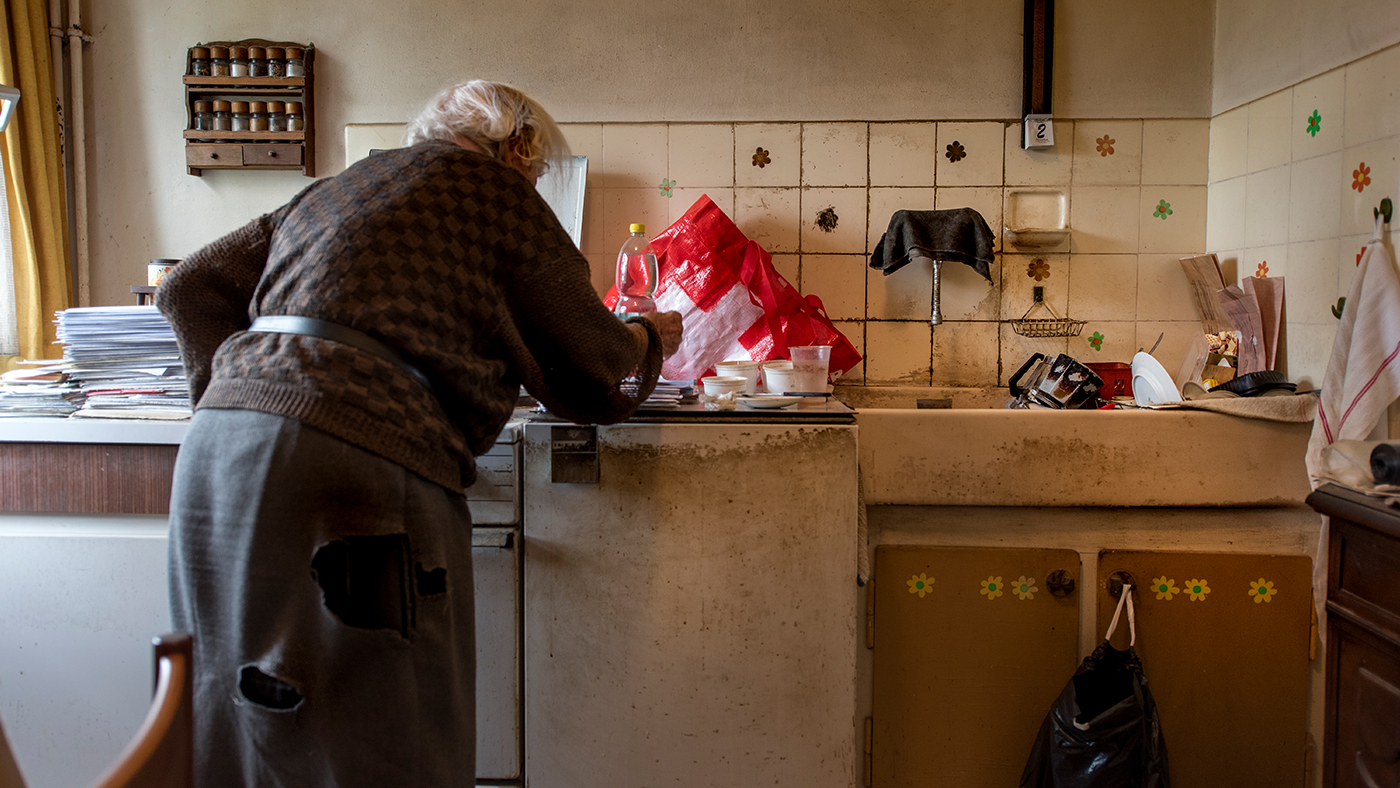 Une personne âgée se tient dans sa petite cuisine et s'occupe de la vaisselle.