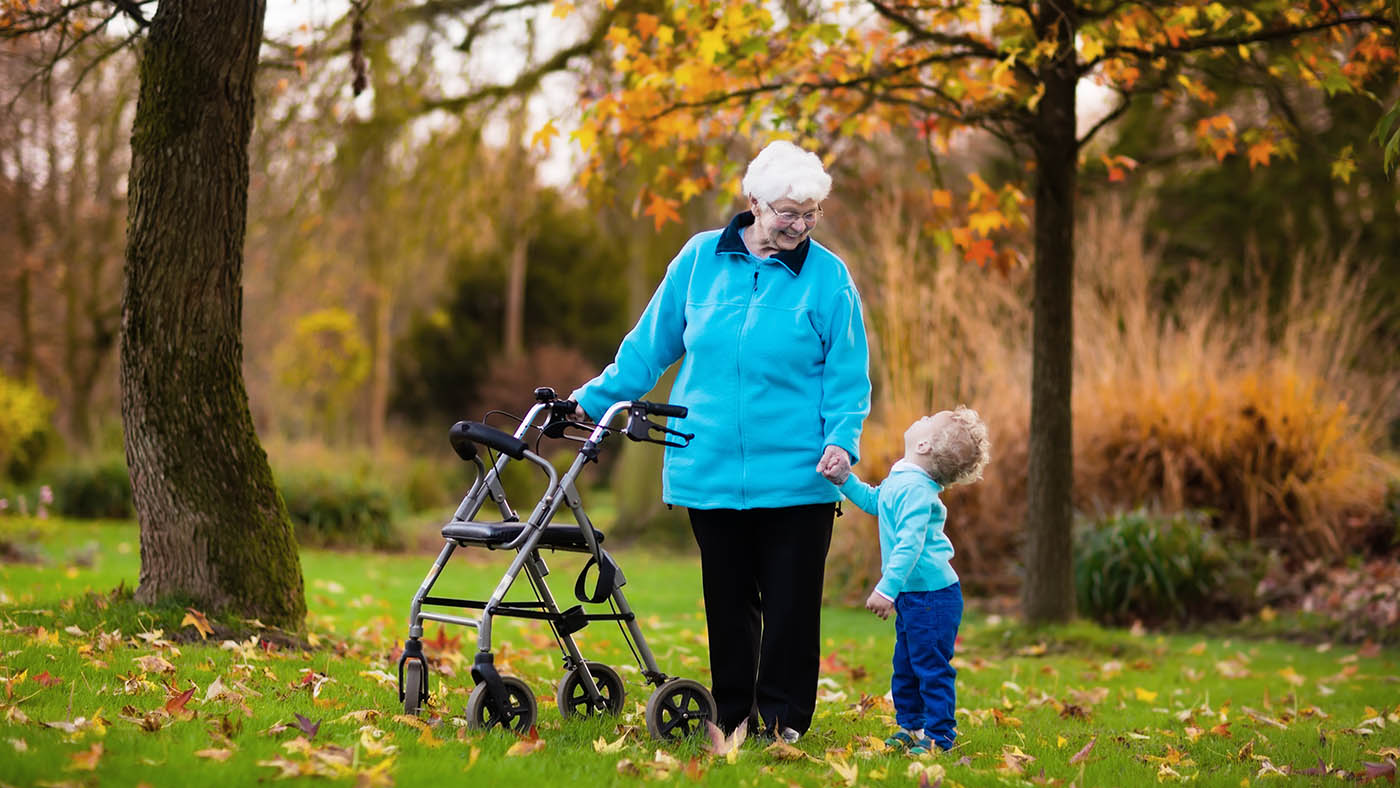 Dans un jardin automnal, une grand-mère en déambulateur tient son petit-fils par la main et lui sourit.