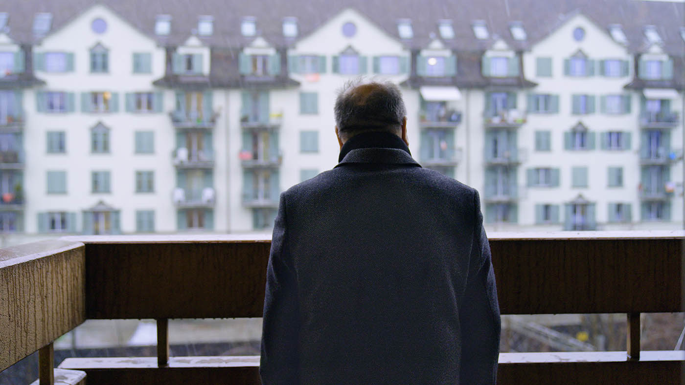 Un homme seul se tient sur un balcon et regarde une rangée d'immeubles.
