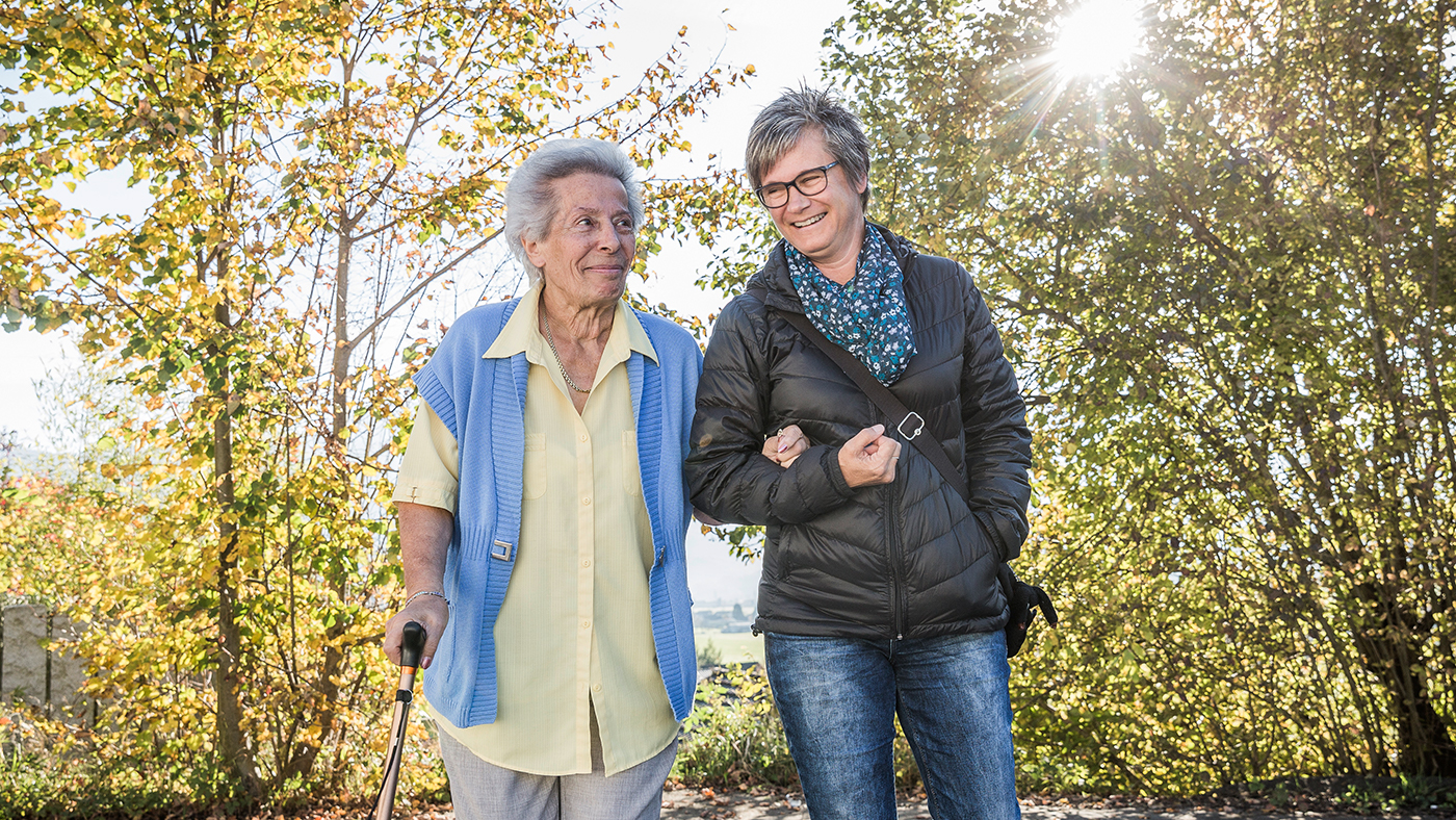 Eine Seniorin spaziert mit ihrer Betreuerin im Wald. 